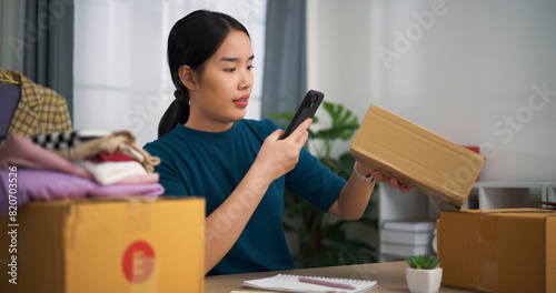 Portrait of Young asian woman business owner sitting at desk use smartphone scan QR code on boxes packing for shipping, Online Shopping