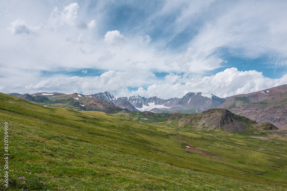 Scenic alpine landscape with green hills and rocks with view to big glacier and large snow-capped mountain range far away under clouds in blue sky in changeable weather. Awesome high snowy mountains. Scenic alpine landscape with green hills and rocks with view to big glacier and large snow-capped mountain range far away under clouds in blue sky in changeable weather. Awesome high snowy mountains.