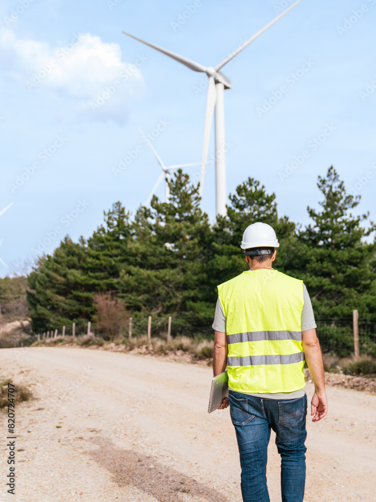 A technician in a hard hat and reflective vest sits on a car trunk working on a laptop near wind turbines on a dirt road. A technician in a hard hat and reflective vest sits on a car trunk working on a laptop near wind turbines on a dirt road.