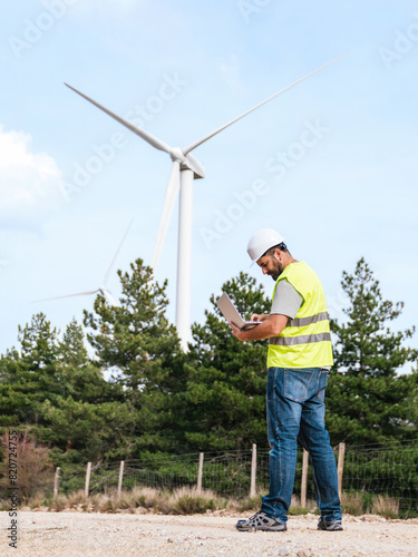 A technician in a high-visibility vest and hard hat uses a laptop while standing in an open field near wind turbines.