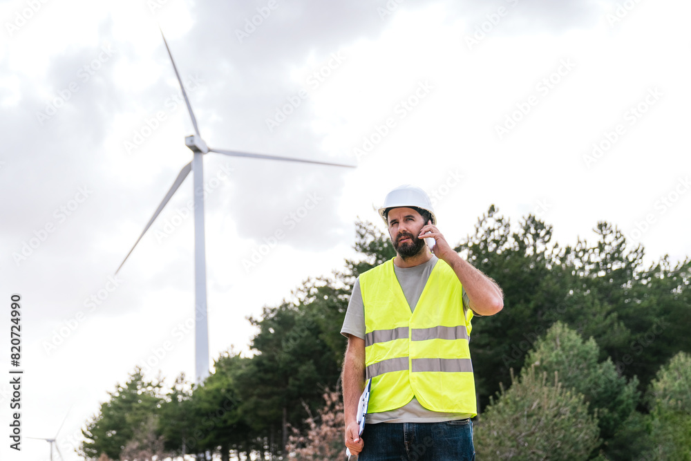 A wind turbine technician speaks on the phone, holding a clipboard, wearing a hard hat and safety vest in a wind farm. A wind turbine technician speaks on the phone, holding a clipboard, wearing a hard hat and safety vest in a wind farm.