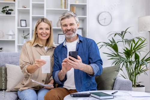 Happy mature couple using a smartphone for online banking while sitting on a sofa at home. They are handling finances together in a cozy living room.