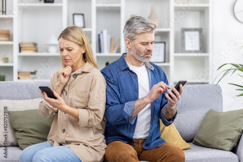 Mature couple sitting back-to-back on a couch using smartphones with serious expressions, both focused on their devices in a modern home interior.