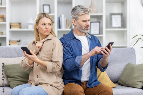 Mature couple sitting back to back on a couch, both looking at their smartphones with unhappy expressions. Concept of relationship issues and technology's impact.