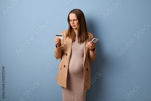 Confused sad upset disappointed pregnant woman wearing dress and jacket isolated over blue background making online payment has not enough money on credit card for purchase