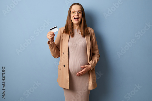 Excited happy cheerful pregnant woman wearing dress and jacket isolated over blue background exclaiming with excitement showing credit card looking at camera choosing new bank