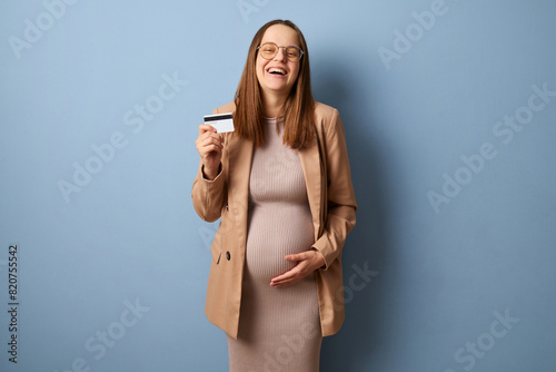 Laughing positive pregnant woman wearing dress and jacket looking at camera while stroking her belly doing banking transaction with credit card isolated over blue background