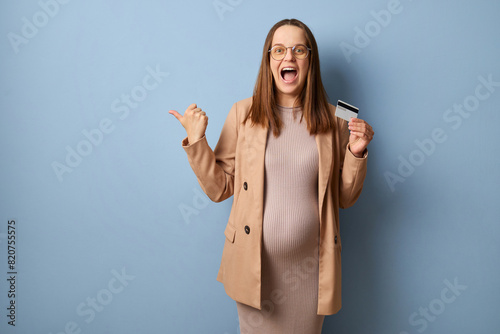 Amazed pregnant woman wearing dress and jacket isolated over blue background making advertisement of bank showing credit card and indicating at empty space for promotional text
