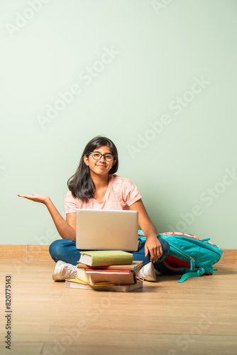 Indian asian intellegent school girl in casual wear using laptop for study sitting on floor isolated pointing or advertising to blank space photo