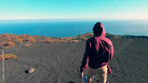 Person dressed in a red hooded sweatshirt walking on a volcanic area with the sea in the background on a sunny and windy day.