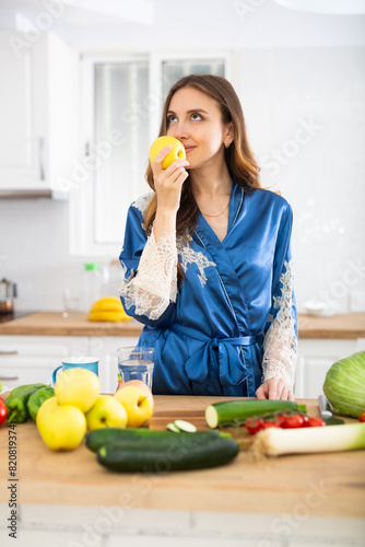 Satisfied woman in dressing gown eating apples in kitchen during breakfast