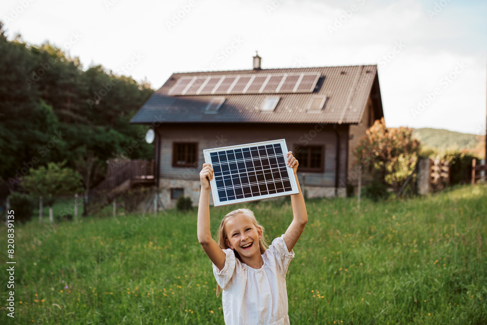 Obraz premium Little girl with model of solar panel, standing in the middle of meadow, house with solar panels behind. Concept of renewable resources.