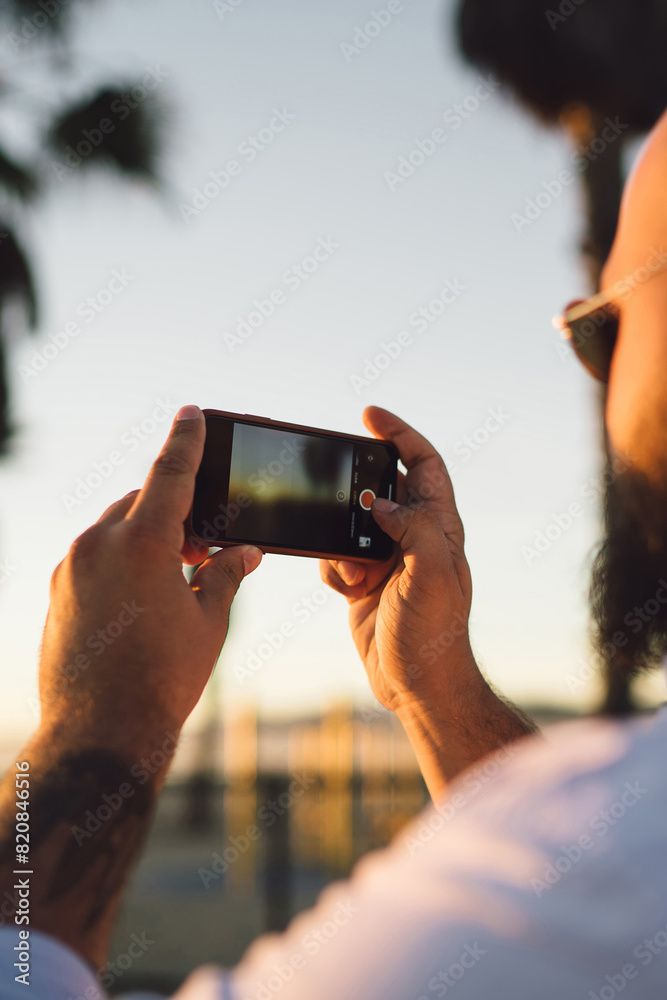 Close up rear view of young male holding in hands smartphone with modern camera for taking photos of beautiful sunset, cropped view of hipster guy shooting evening landscape on mobile telephone