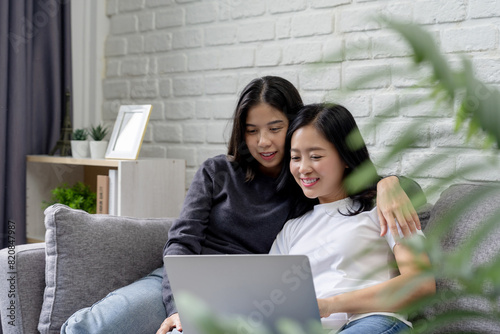 Happy Asian lesbian couple hugging each other having a good time. Together while using a laptop, sitting on a sofa, happy moments, LGBT pride