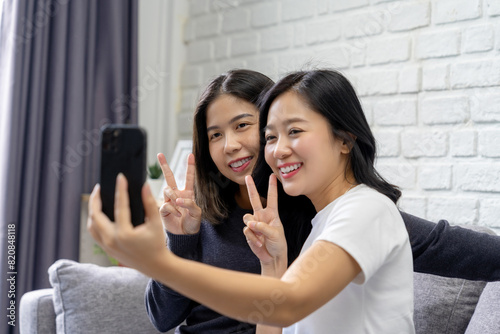 Two Asian female friends taking pictures together sitting on a sofa in a house Showing love, happy moments, LGBT pride