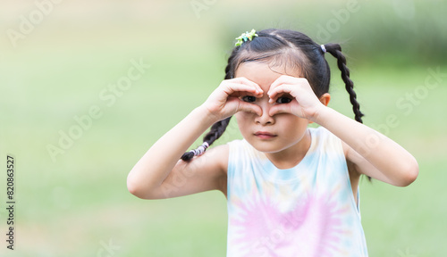 Portrait of Asian child girl in park playing outdoor in nature. Happy little active girl using hands gesture binocular, smiling in casual clothes. Education, healthy activities