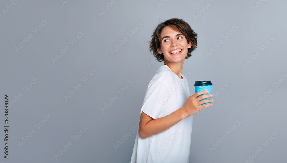 Close up photo of beautiful modern woman with trendy hairstyle in white t-shirt smiling and holding takeaway coffee in paper cup, isolated over grey background. Close up photo of beautiful modern woman with trendy hairstyle in white t-shirt smiling and holding takeaway coffee in paper cup, isolated over grey background.