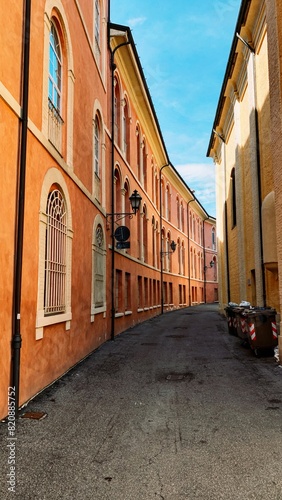 Narrow alley between historic buildings with arched windows and orange walls on a sunny day. Trash bins lined up on the side, blue sky in the background.