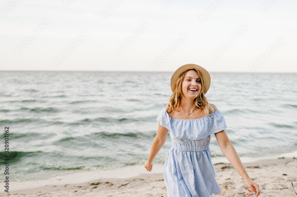 Young attractive smiling blond woman in straw hat joyfully walks near sea. Happy beautiful girl standing on beach ocean and enjoying sunny summer day on vacation. Portrait of stylish female on sand. Young attractive smiling blond woman in straw hat joyfully walks near sea. Happy beautiful girl standing on beach ocean and enjoying sunny summer day on vacation. Portrait of stylish female on sand.