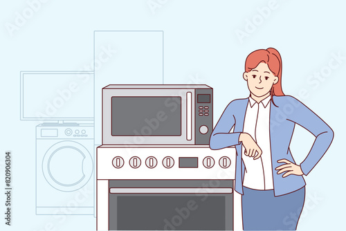 Woman selling household appliances stands near oven and washing machine and looks at camera. Girl smiles and rejoices in purchasing new home appliances with IOT wifi synchronization technology