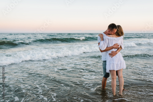 Couple in love hugging and kissing on seashore in sea. Male kisses and hugs female standing on water with big waves ocean and enjoying a summer day. Man embraces woman walking on beach sand sea.