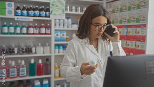 A young attractive hispanic brunette woman works in an indoor pharmacy wearing a lab coat, holding a medicine bottle while talking on the phone and looking at a computer screen.