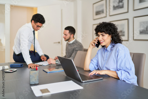 A woman is talking on her cell phone while sitting at a desk with a laptop