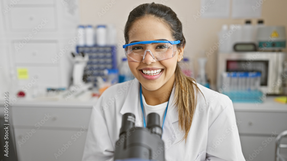 A smiling young hispanic woman in a lab coat and safety glasses works in a laboratory setting with a microscope in the forefront. A smiling young hispanic woman in a lab coat and safety glasses works in a laboratory setting with a microscope in the forefront.