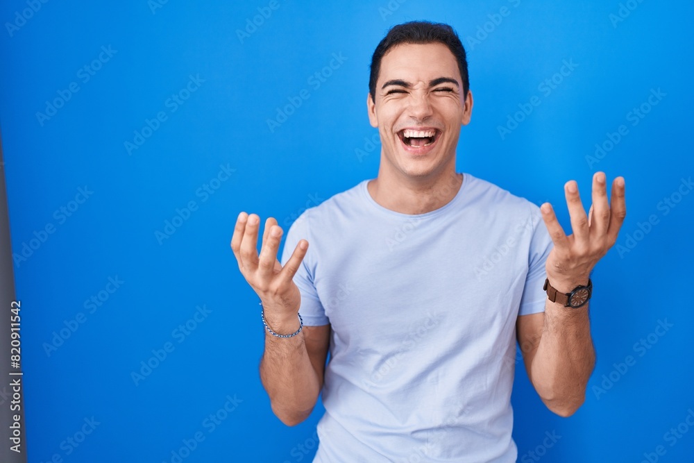 Young hispanic man standing over blue background celebrating mad and crazy for success with arms raised and closed eyes screaming excited. winner concept