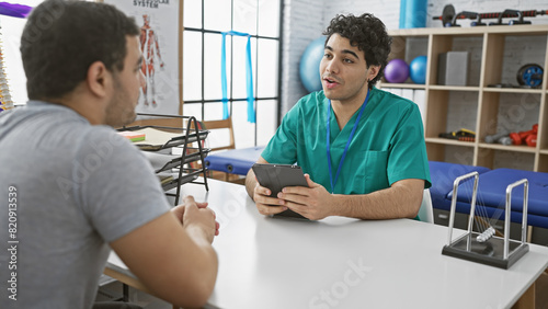 A male therapist in scrubs discusses treatment with a patient in a bright physiotherapy clinic. photo