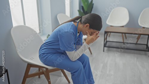 A distressed young woman in blue scrubs sits in a bright hospital room, covering her face with her hands. © Krakenimages.com