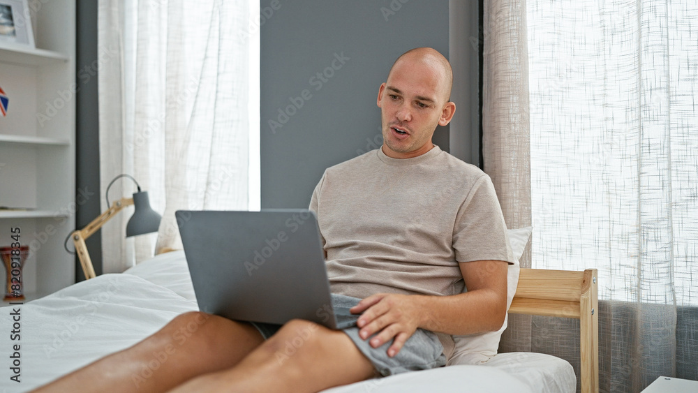 Young hispanic man doing video call with laptop sitting on the bed at bedroom