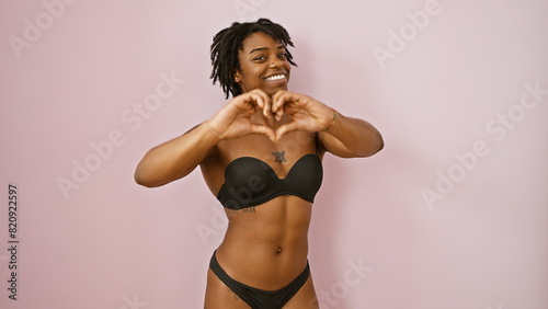 A cheerful young woman with dreadlocks makes a heart gesture against a pink isolated background.