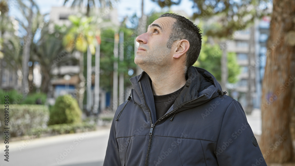 Hispanic man in contemplative pose stands outdoors in a city park, conveying a sense of reflection.