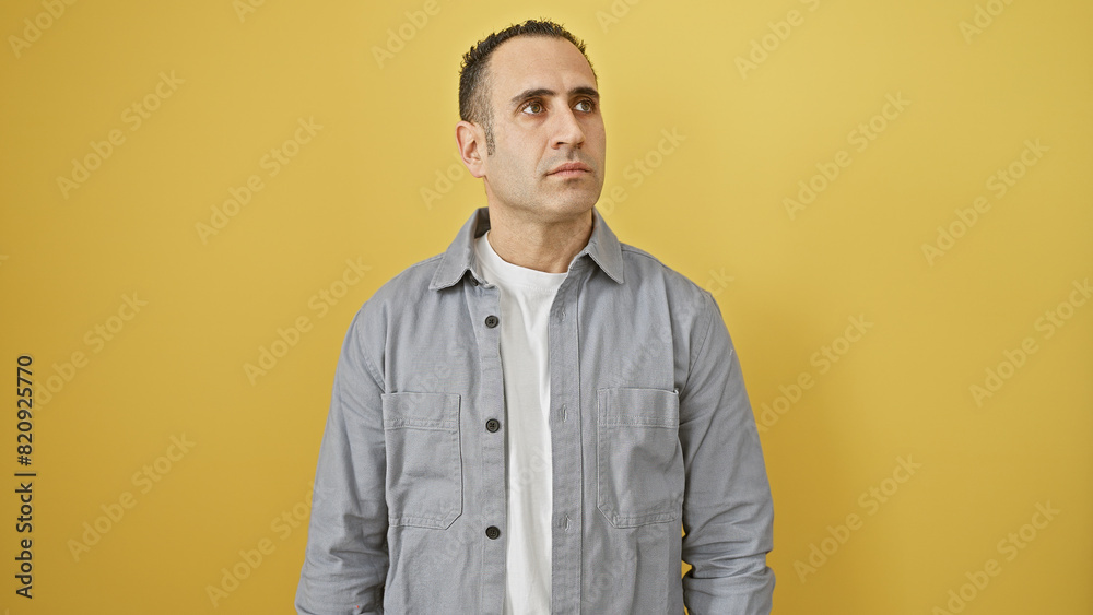 Hispanic man in grey shirt posing against an isolated yellow background looking away thoughtfully. Hispanic man in grey shirt posing against an isolated yellow background looking away thoughtfully.