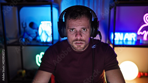 Concerned hispanic man with headphones in a neon-lit gaming room at night  emanating intensity and focus.