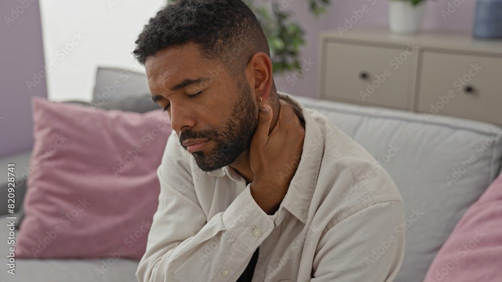 A young african american man with a beard feeling neck pain while sitting on a couch indoors. A young african american man with a beard feeling neck pain while sitting on a couch indoors.