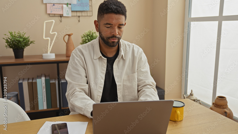 A focused african american man with a beard works on a laptop in a modernly decorated home office. A focused african american man with a beard works on a laptop in a modernly decorated home office.