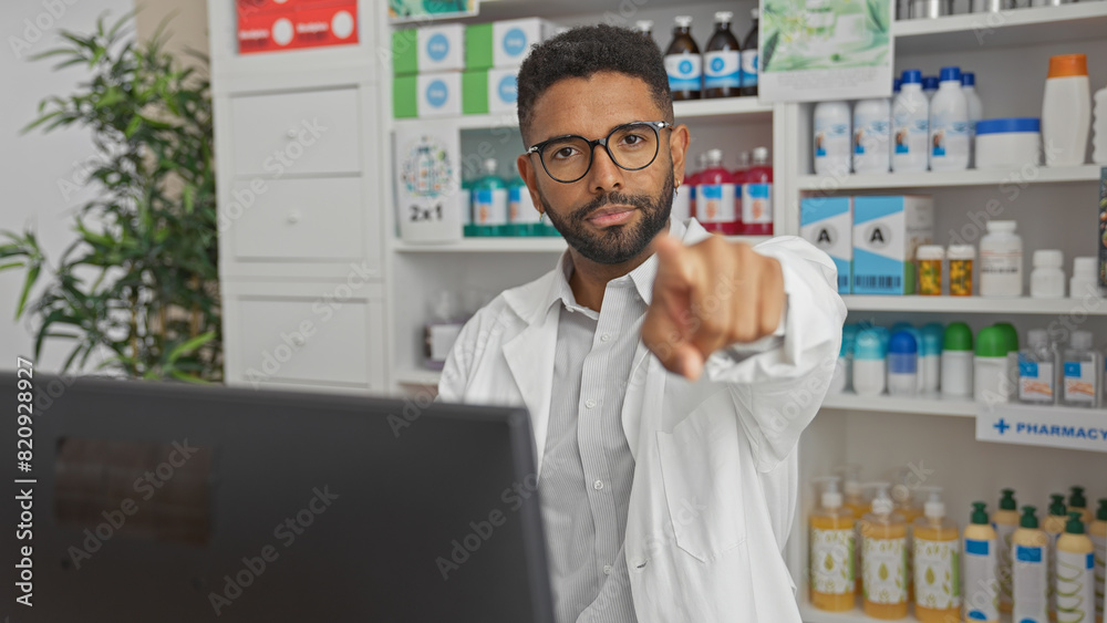 A confident young male pharmacist points towards the camera in a well-stocked, modern drugstore
