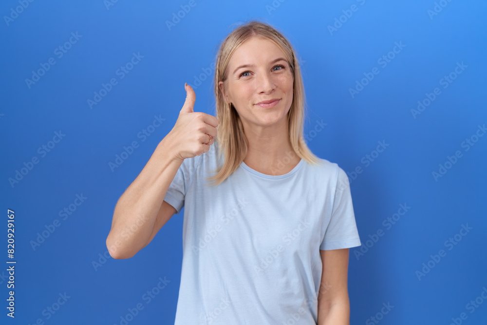Young caucasian woman wearing casual blue t shirt doing happy thumbs up gesture with hand. approving expression looking at the camera showing success. Young caucasian woman wearing casual blue t shirt doing happy thumbs up gesture with hand. approving expression looking at the camera showing success.