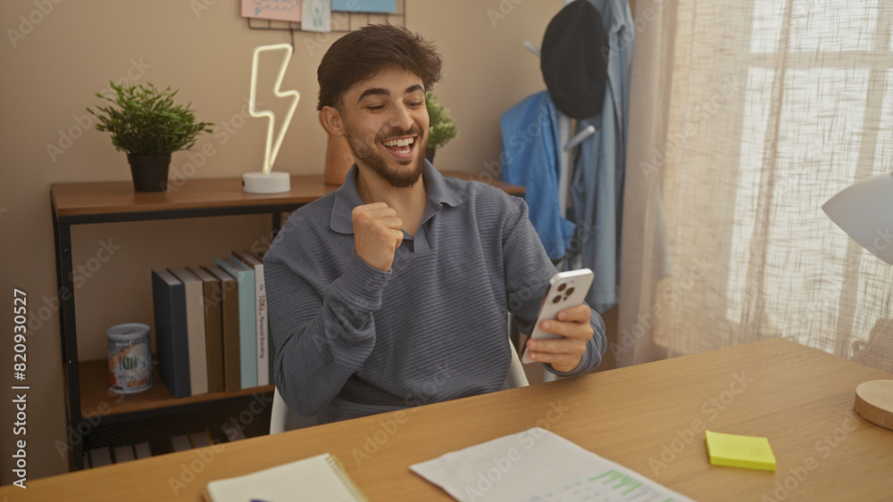 A cheerful young man with a beard celebrates a victory on his smartphone in a cozy living room. A cheerful young man with a beard celebrates a victory on his smartphone in a cozy living room.