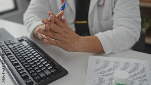 A hispanic man working indoors at a clinic with a computer, papers, and pens, portraying a professional healthcare environment.