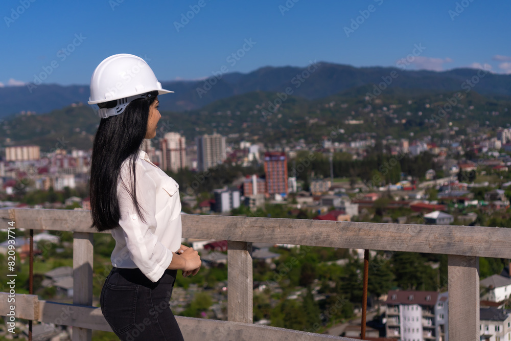 Young brunette resident architect in a hard hat on a construction site against the background of the city and mountains on a sunny day. Successful inspector. Young brunette resident architect in a hard hat on a construction site against the background of the city and mountains on a sunny day. Successful inspector.