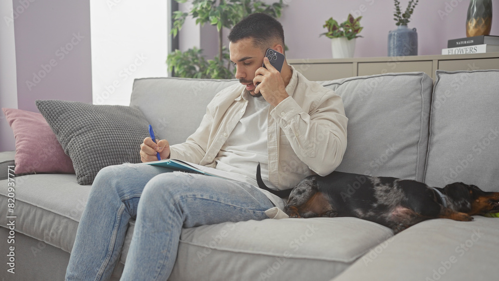A young hispanic man multitasking with a phone call and notetaking, alongside a resting dachshund on a couch in a cozy living room.