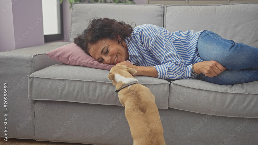 A smiling middle-aged woman enjoys bonding with her tan chihuahua on a grey sofa indoors, depicting a cozy home setting. A smiling middle-aged woman enjoys bonding with her tan chihuahua on a grey sofa indoors, depicting a cozy home setting.