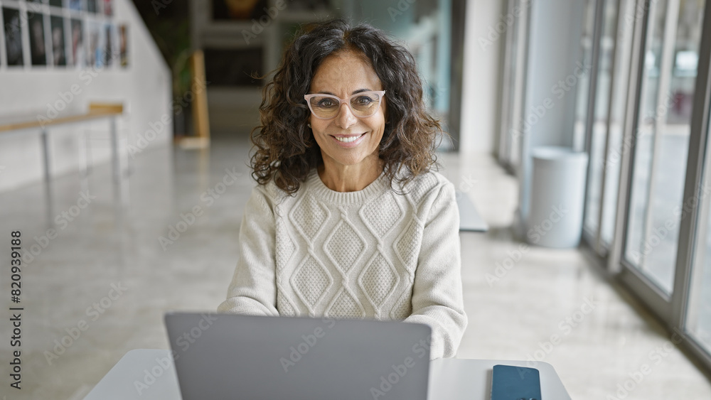 A smiling hispanic woman in a modern office wearing glasses while working on a laptop. A smiling hispanic woman in a modern office wearing glasses while working on a laptop.