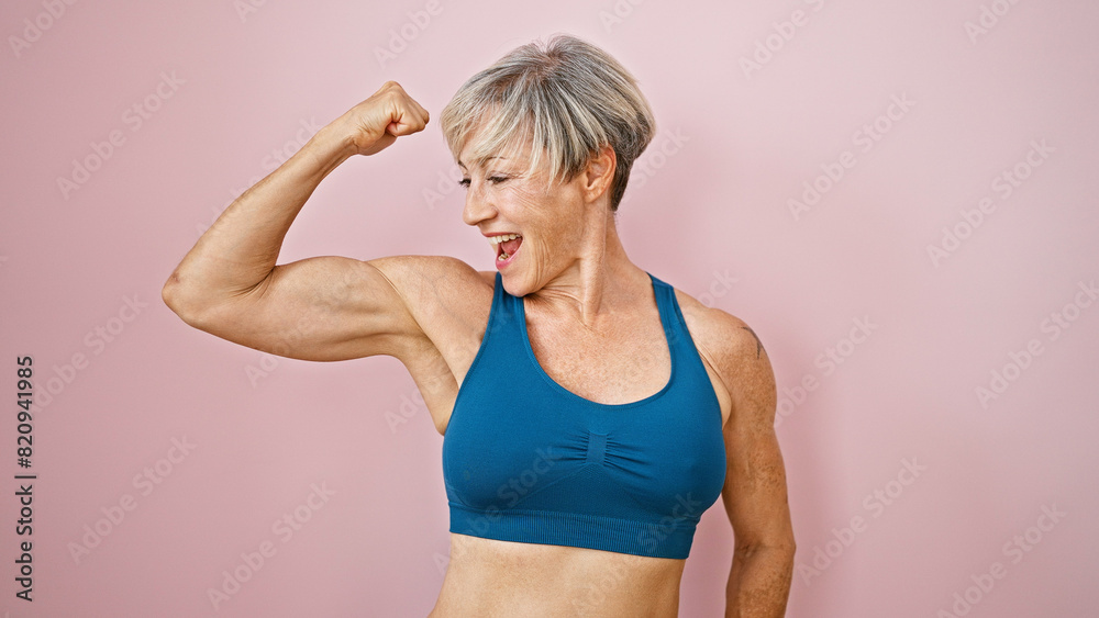A fit, mature woman with short grey hair flexes her muscles against a pink isolated background, displaying strength and vitality. A fit, mature woman with short grey hair flexes her muscles against a pink isolated background, displaying strength and vitality.