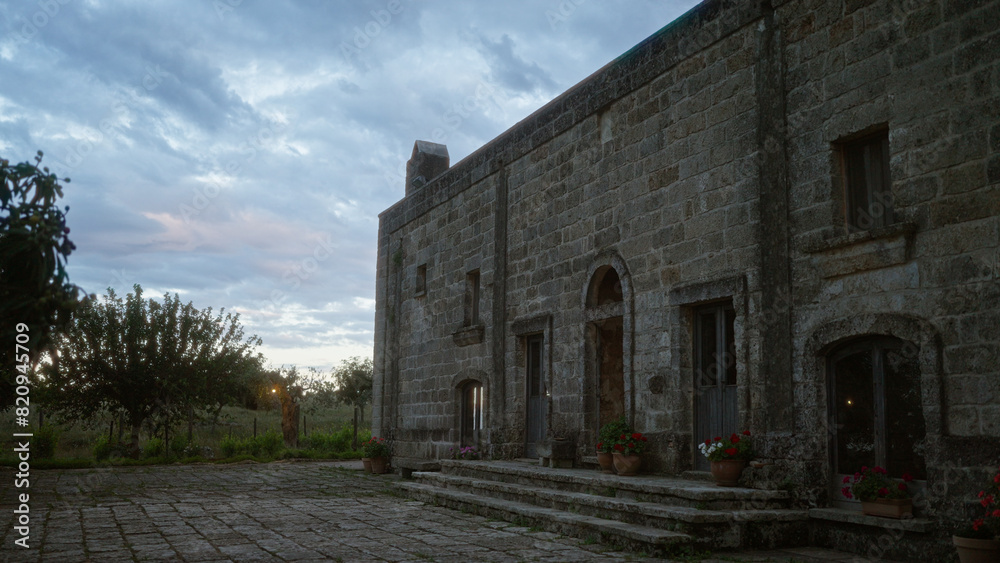 Stone building in puglia, italy, at dusk with potted flowers and a serene sky, surrounded by lush greenery.