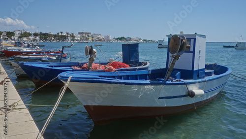 Fishing boats docked at the harbor in porto cesareo  italy  with a clear blue sky and coastal town in the background.