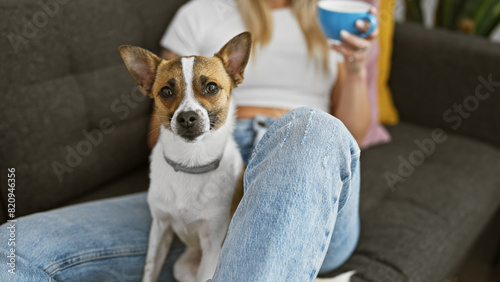 A woman holding a mug relaxes with her small dog on a cozy couch indoors.